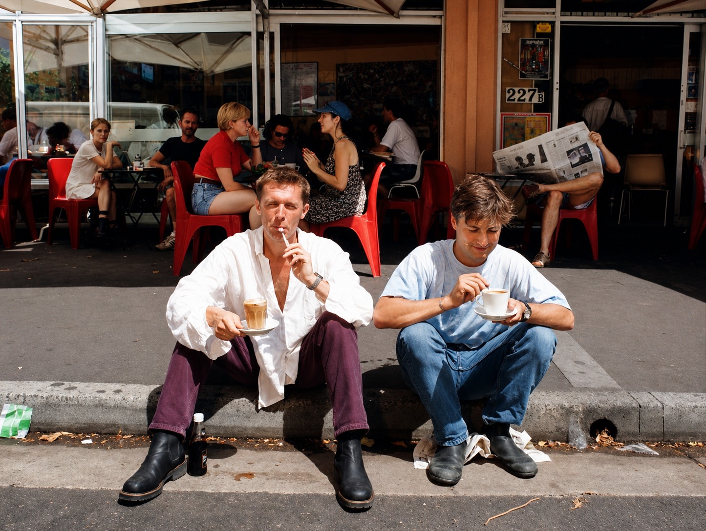 Three young men sitting on the kerb outside Tropicana Caffe in the late 1980s, the original two-room frontage and awning visible behind them
